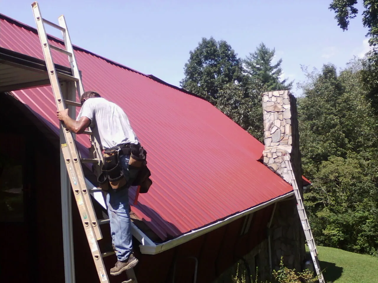 Roofer on a ladder beside a red metal roof and stone chimney — Soffit & Fascia Repair by Roofing Brothers in Machesney Park